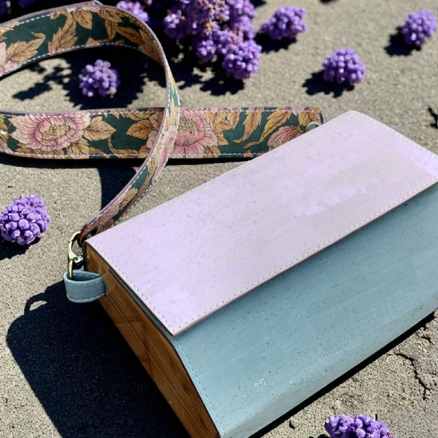 Two handbags, one with a floral strap, on a sandy surface with purple flowers.