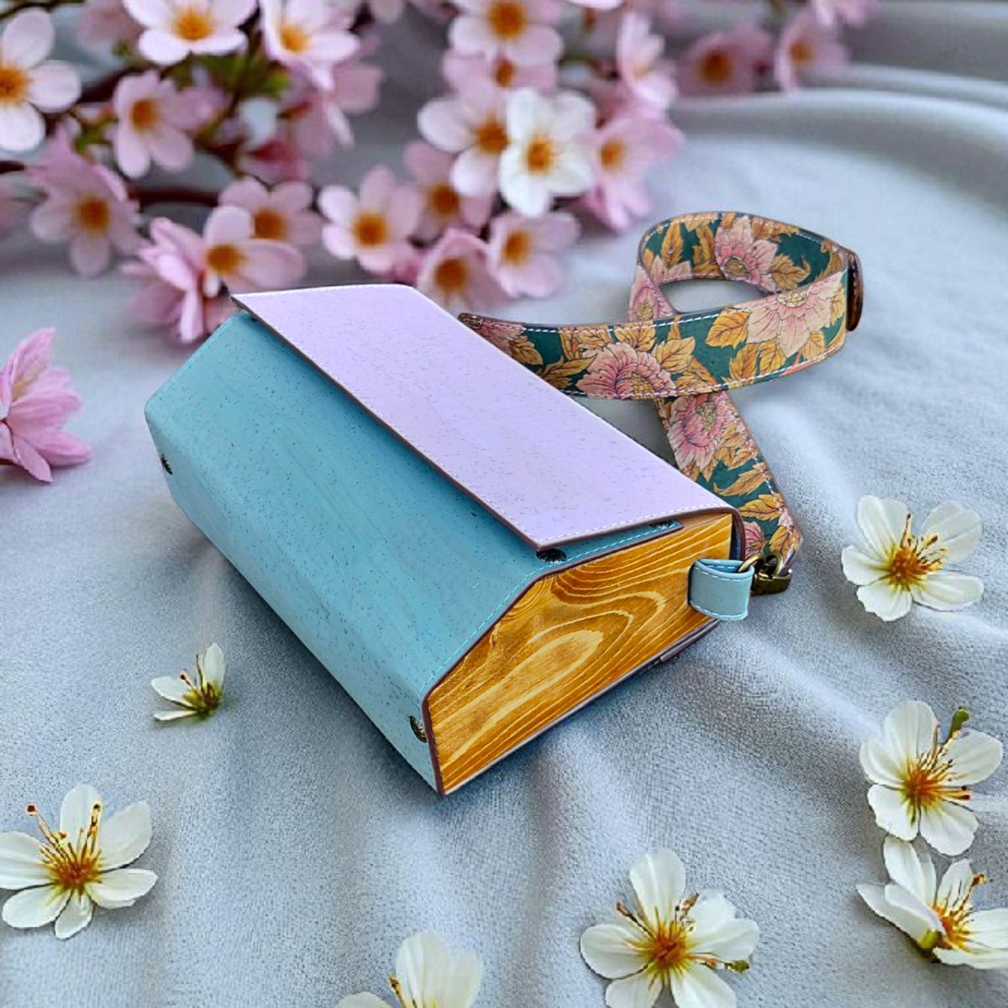 Blue notebook with a pink cover and floral strap, surrounded by flowers on a light surface.
